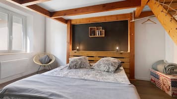 A bedroom with a wooden headboard, a bed with patterned bedding, a chair, and a small shelf with books.