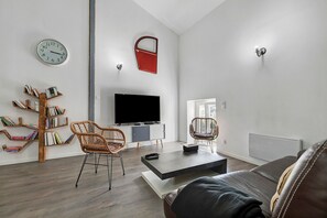 Een moderne woonkamer met een flatscreen televisie, een boekenkast, twee rieten stoelen, een salontafel, en een rode tas aan de wand.