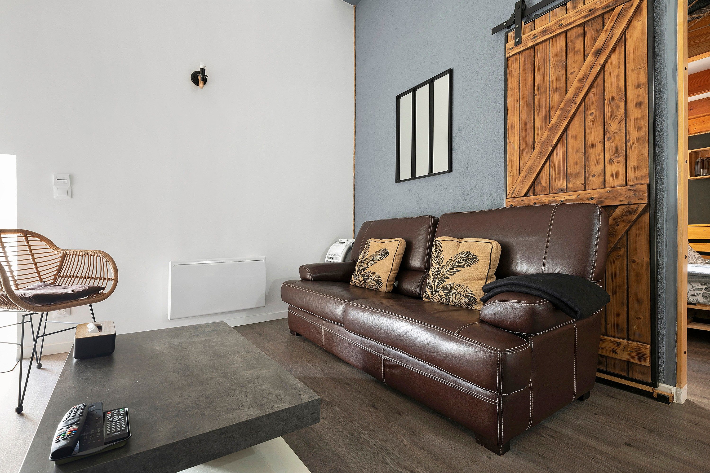 Un canapé en cuir marron avec deux coussins, une chaise en osier, une petite table avec une télécommande et une porte en bois avec une poignée en métal.