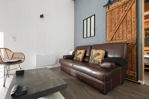 Un canapé en cuir marron avec deux coussins, une chaise en osier, une petite table avec une télécommande et une porte en bois avec une poignée en métal.