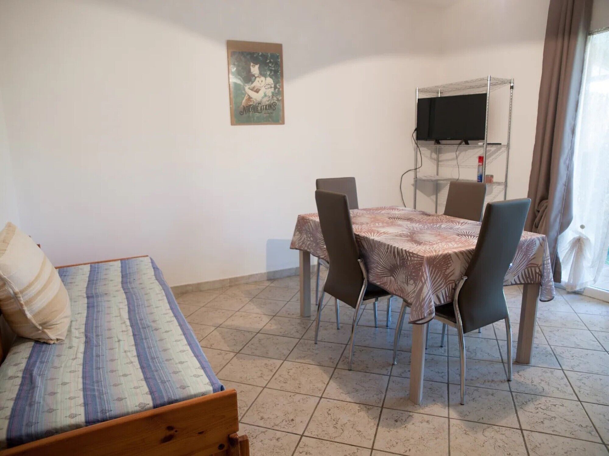 Una habitación con cama, una mesa de comedor con sillas, un televisor sobre un mueble bajo y un ventanal con cortinas.