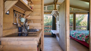 Interno di una baita in legno con angolo cottura, un letto con copriletto floreale e vista sul verde circostante.