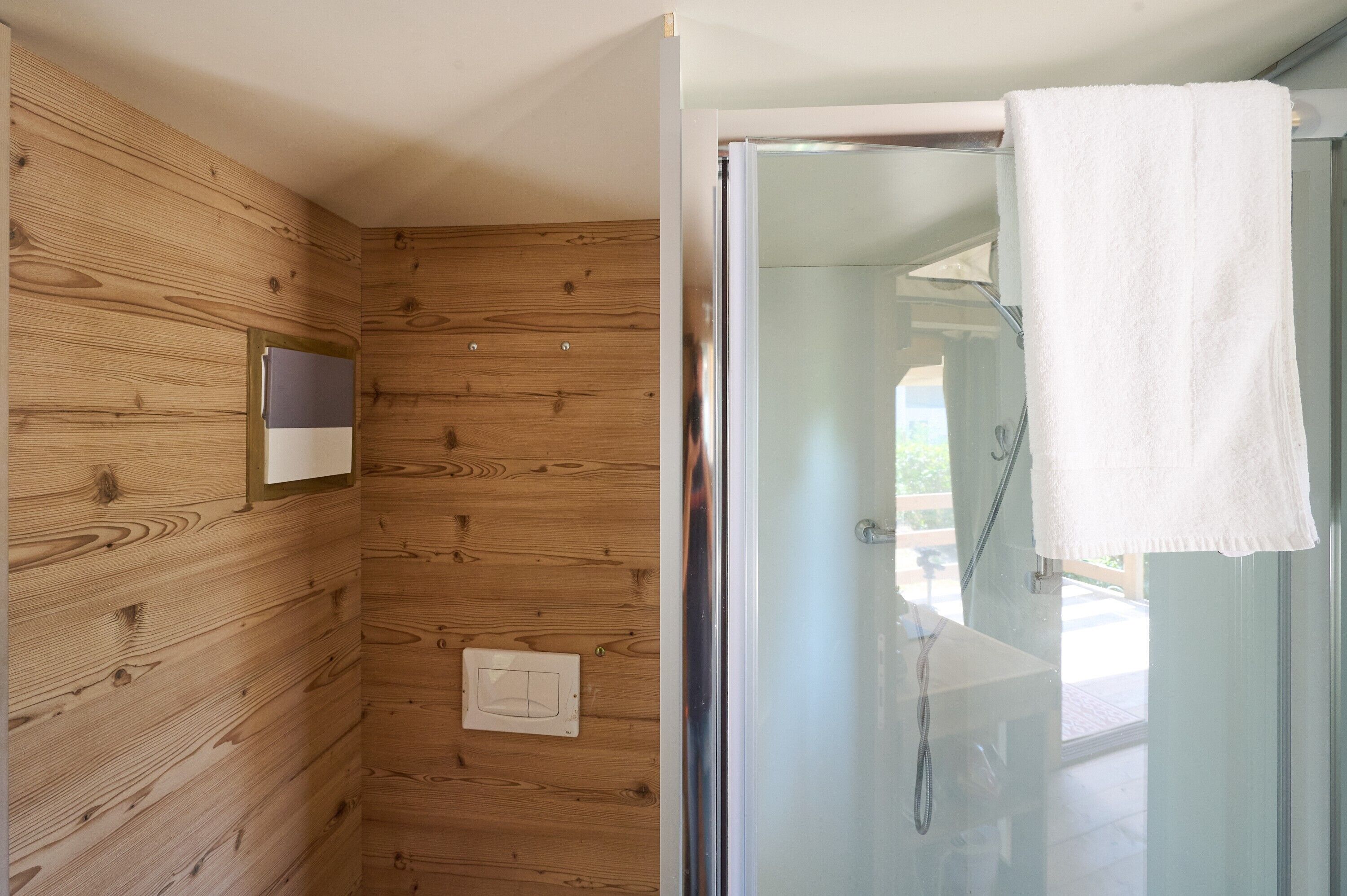 A wooden-paneled bathroom with a glass shower enclosure and a white towel hanging on the door.