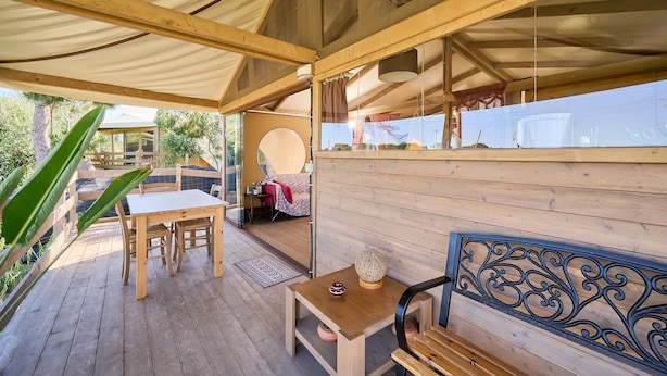 Une terrasse en bois avec une table et des chaises, un banc et une vue sur un bâtiment, visible à travers une porte vitrée.