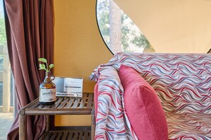 A cozy room with a bed, a small table with a bottle and a radio, and a window with curtains.