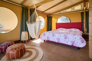 Une chambre à coucher avec un lit, un fauteuil suspendu, des poufs et de grandes fenêtres.