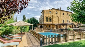 A poolside area with lounge chairs, a wooden fence, and a building with arched windows. - Farmhouse 'Mas Alba' with Mountain View, Private Pool and Wi-Fi (Terradelles Girona)