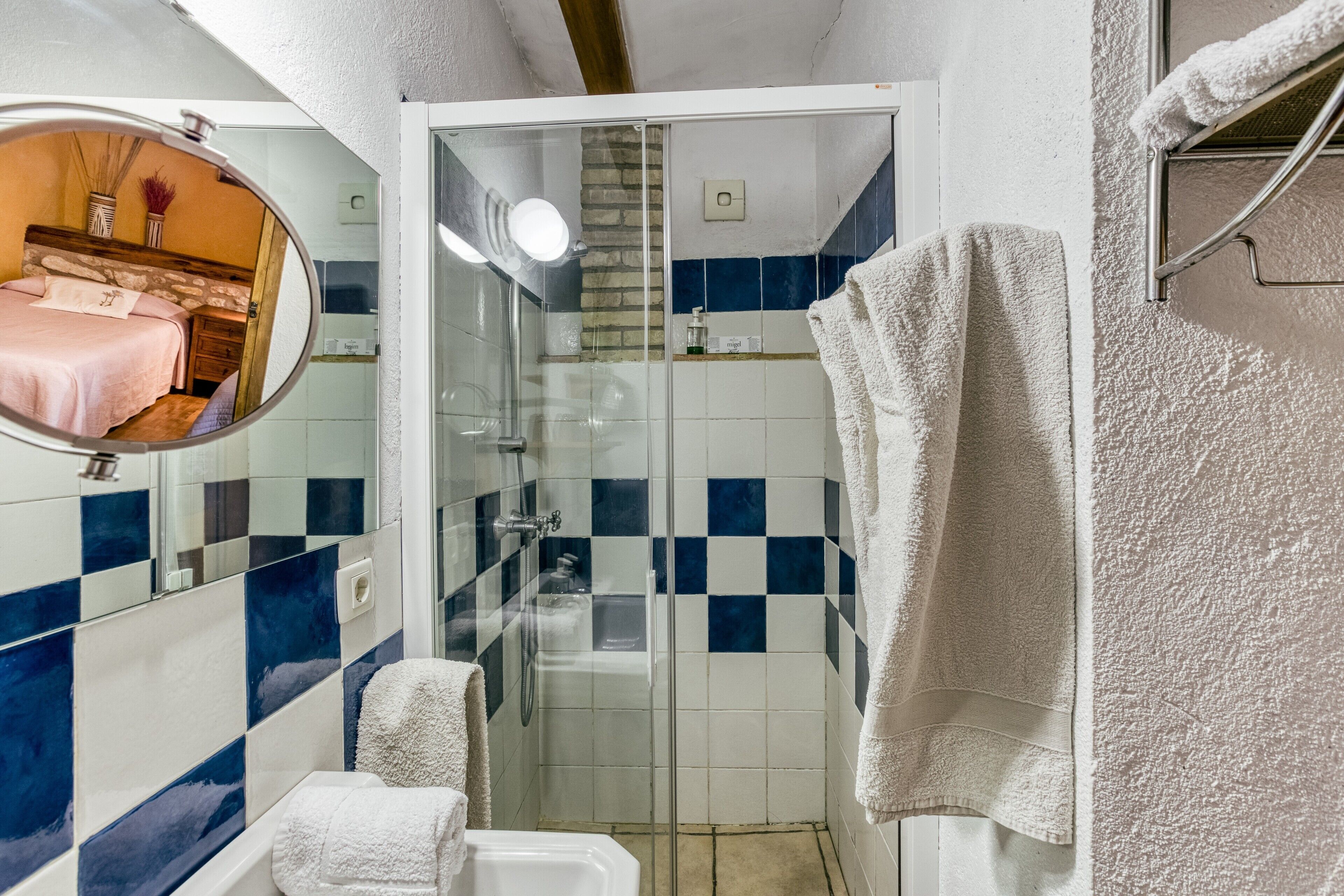 A bathroom with a glass shower enclosure, white bathtub, and a mirror reflecting a bedroom.