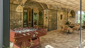 A covered outdoor area with a foosball table, wicker chairs, and a stone wall with a door. - Farmhouse 'Mas Alba' with Mountain View, Private Pool and Wi-Fi (Terradelles Girona)
