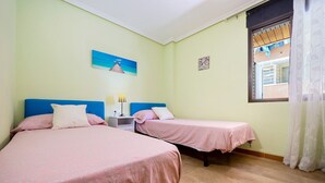 A room with two beds, a nightstand, a lamp, a window with a curtain, and a wall with a painting. - Apartment 'Salou Centro' with Private Terrace (Salou)