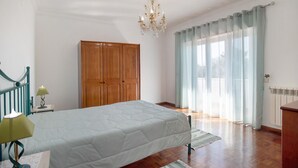 Une chambre à coucher avec un lit, une armoire en bois, un lustre et une vue par une fenêtre avec des rideaux.