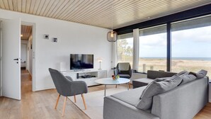 Uma sala de estar moderna com uma televisão grande, sofá cinzento e vista para a praia.