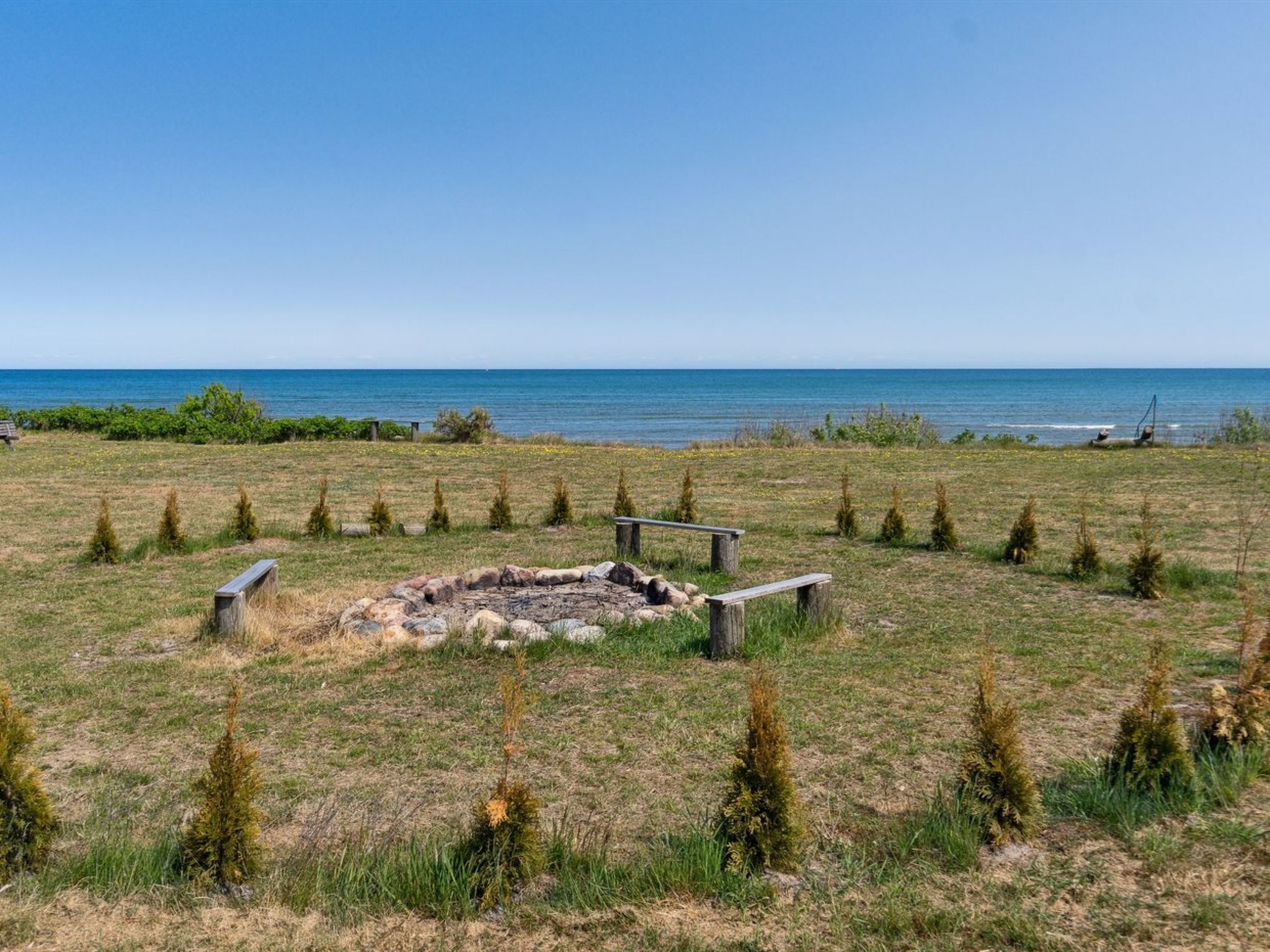 A circular stone structure surrounded by small trees in a grassy field with a view of the sea.