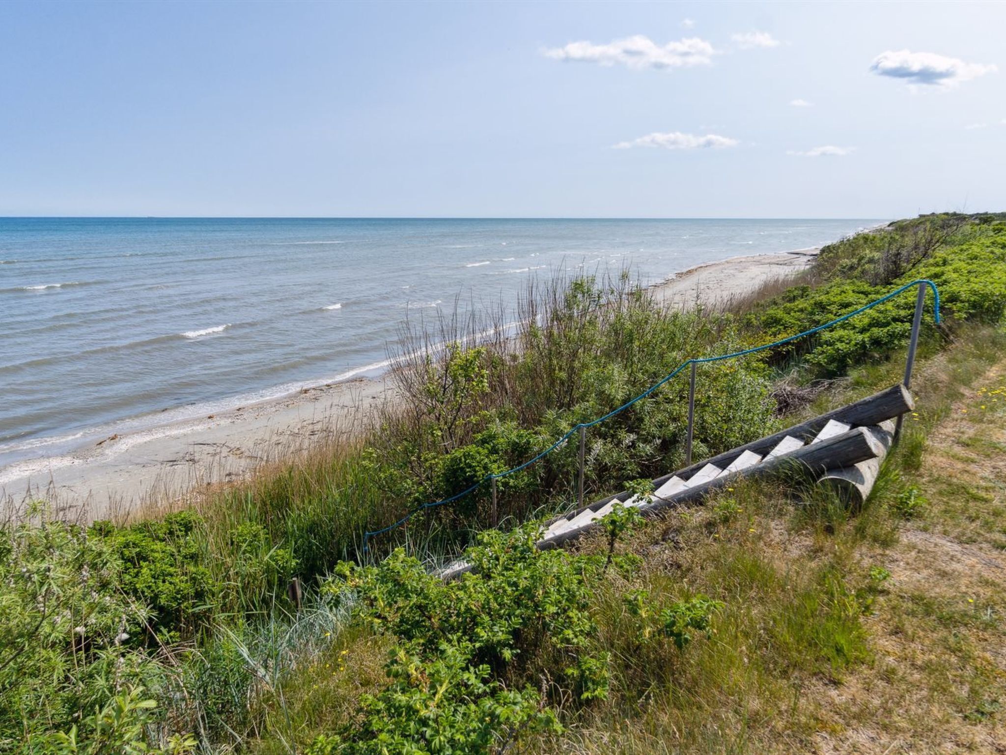 Ein Küstenpfad mit Metallgeländer, der zu einem Strand mit sanften Wellen führt.