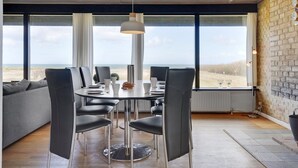 Uma sala de jantar moderna com uma mesa redonda, cadeiras pretas e uma luminária pendente. Janelas amplas oferecem vista para a praia.