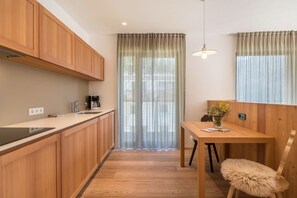 A modern kitchen with wooden cabinets, a dining table with a chair, and a window with sheer curtains.