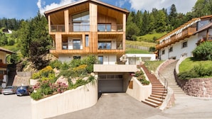 A multi-story wooden house with a garage, surrounded by greenery and a hillside. - Vista Dolomites Apartment "Harmony" with Mountain View, Private Garden & Wi-Fi (S. Cristina)
