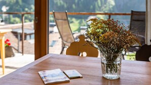 A wooden table with a clear glass vase containing flowers, two postcards, and a view of a mountainous landscape through a glass door. - Vista Dolomites Apartment "Harmony" with Mountain View, Private Garden & Wi-Fi (S. Cristina)