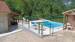 A swimming pool with a fenced deck, surrounded by trees and a small building with a tiled roof.
