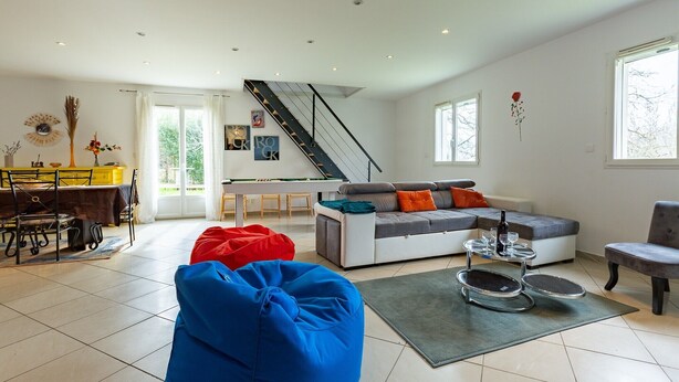 A modern living room with a sofa, a dining area with a table and chairs, a staircase, and a view of the outdoors.
