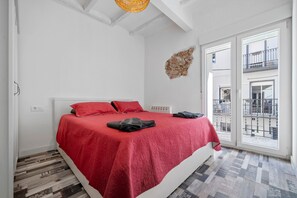 Un dormitorio con colcha roja, cabecera blanca, pequeña decoración en la pared y vista al exterior por un ventanal.