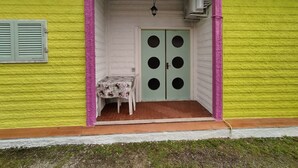 Un edificio con fachada verde, una puerta blanca con seis ventanas circulares, una pequeña mesa con mantel floral y un farol colgante.