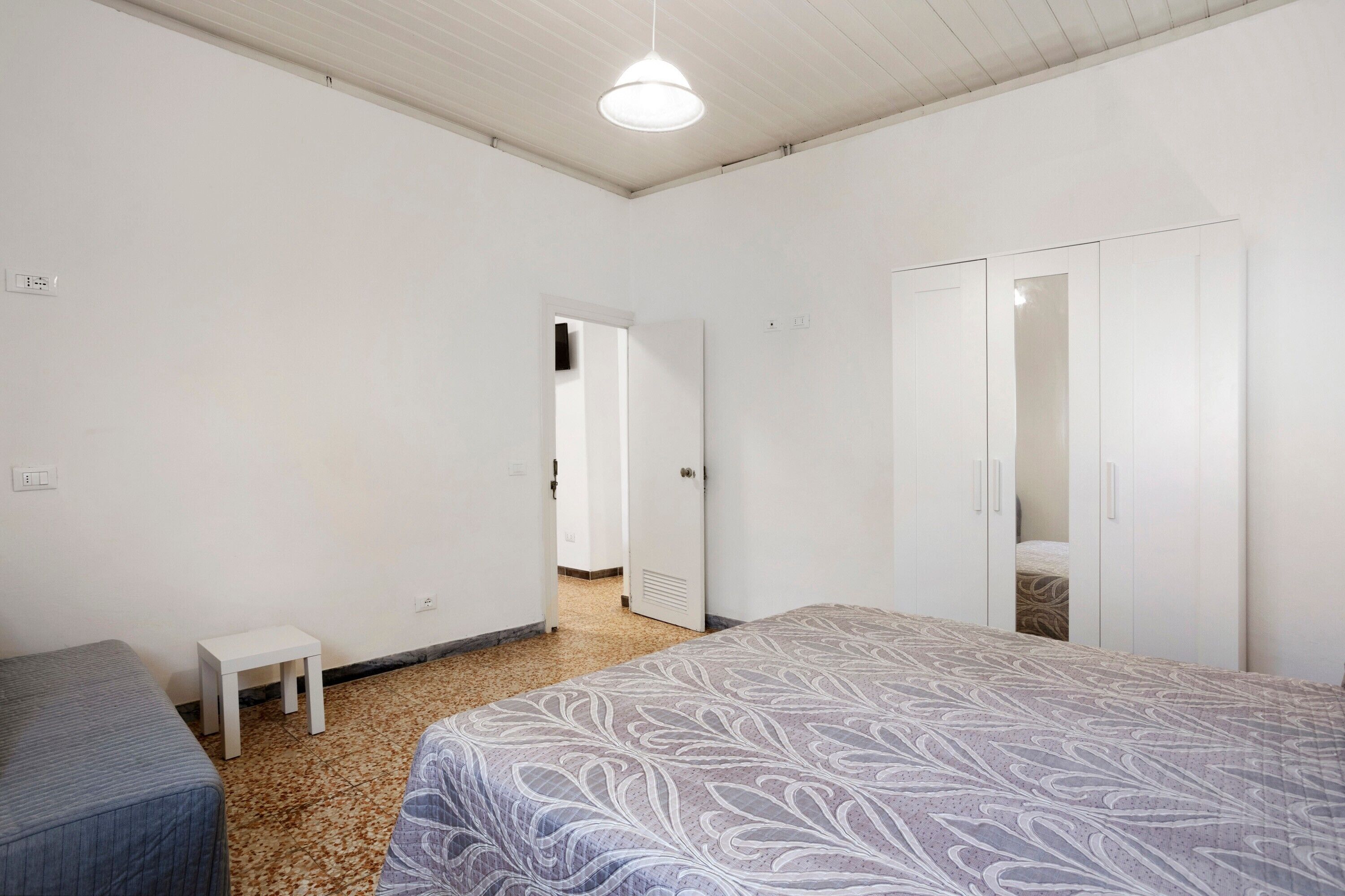 Un dormitorio con una cama, una mesita blanca, un armario y una puerta que da a otra habitación.