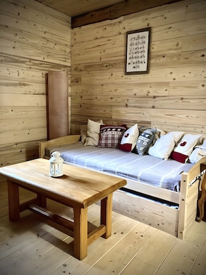 Interior de uma cabana de madeira com uma cama, almofadas, uma mesa e um candeeiro.