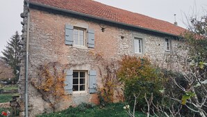 Un edificio de piedra con tejido rojo, dos ventanas con contraventanas y un jardín con césped y arbustos.