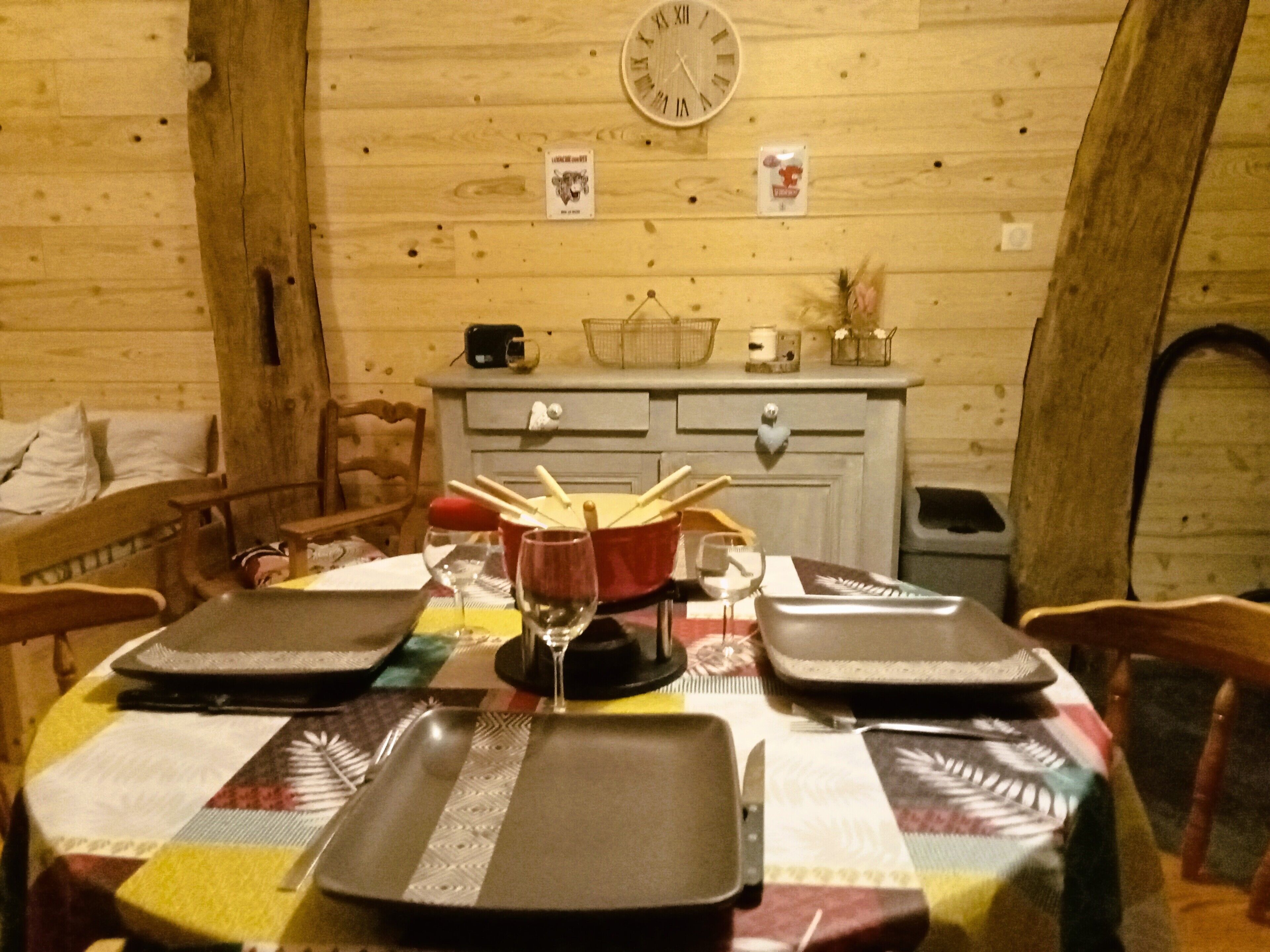 Een houten interieur met een eettafel gedekt voor vier personen, een klok aan de muur, en een keuken met kasten en een spoelbak.