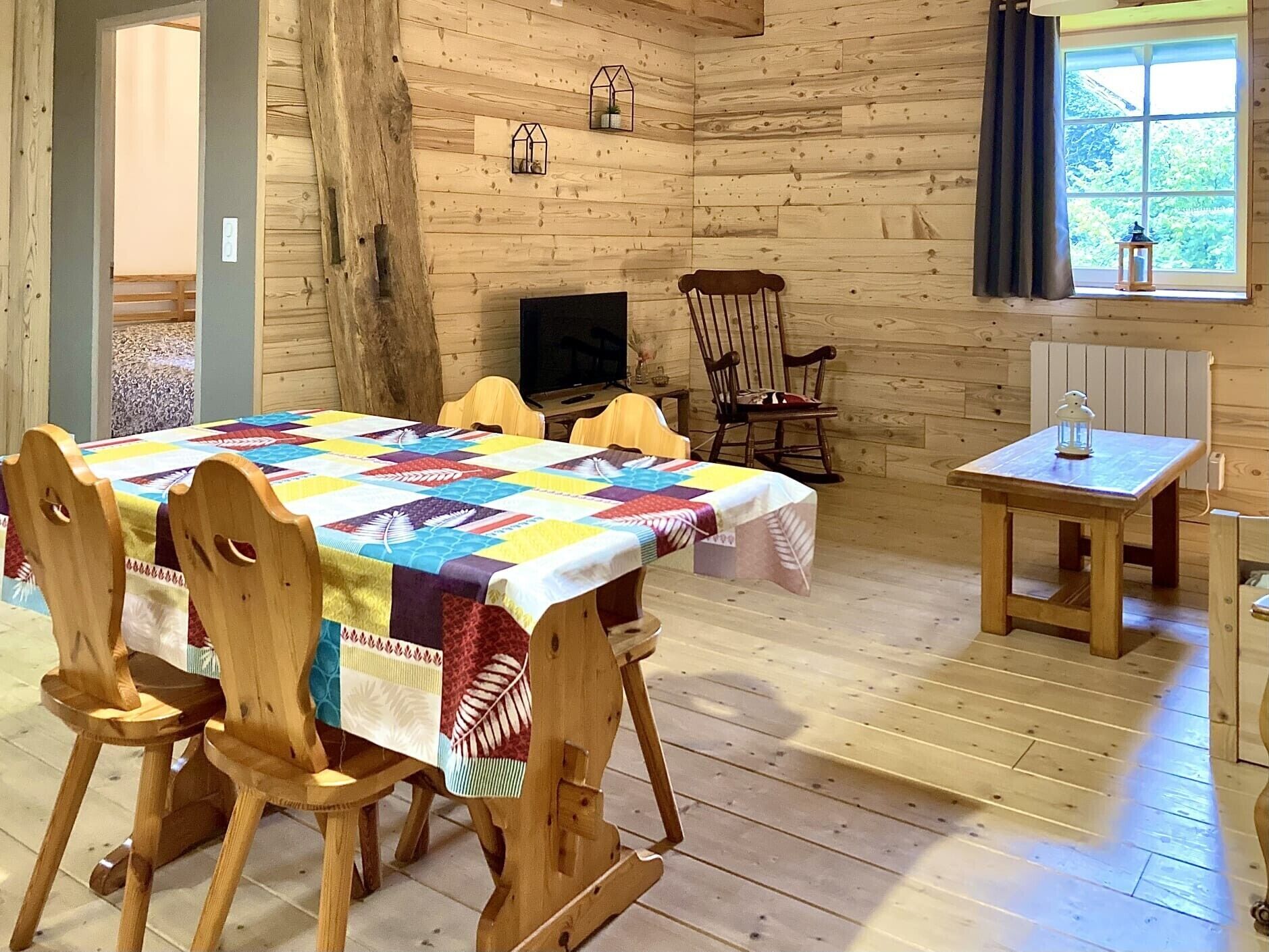 A wooden cabin interior with a dining table, chairs, a television, a rocking chair, a small table with a lantern, and a window with curtains.