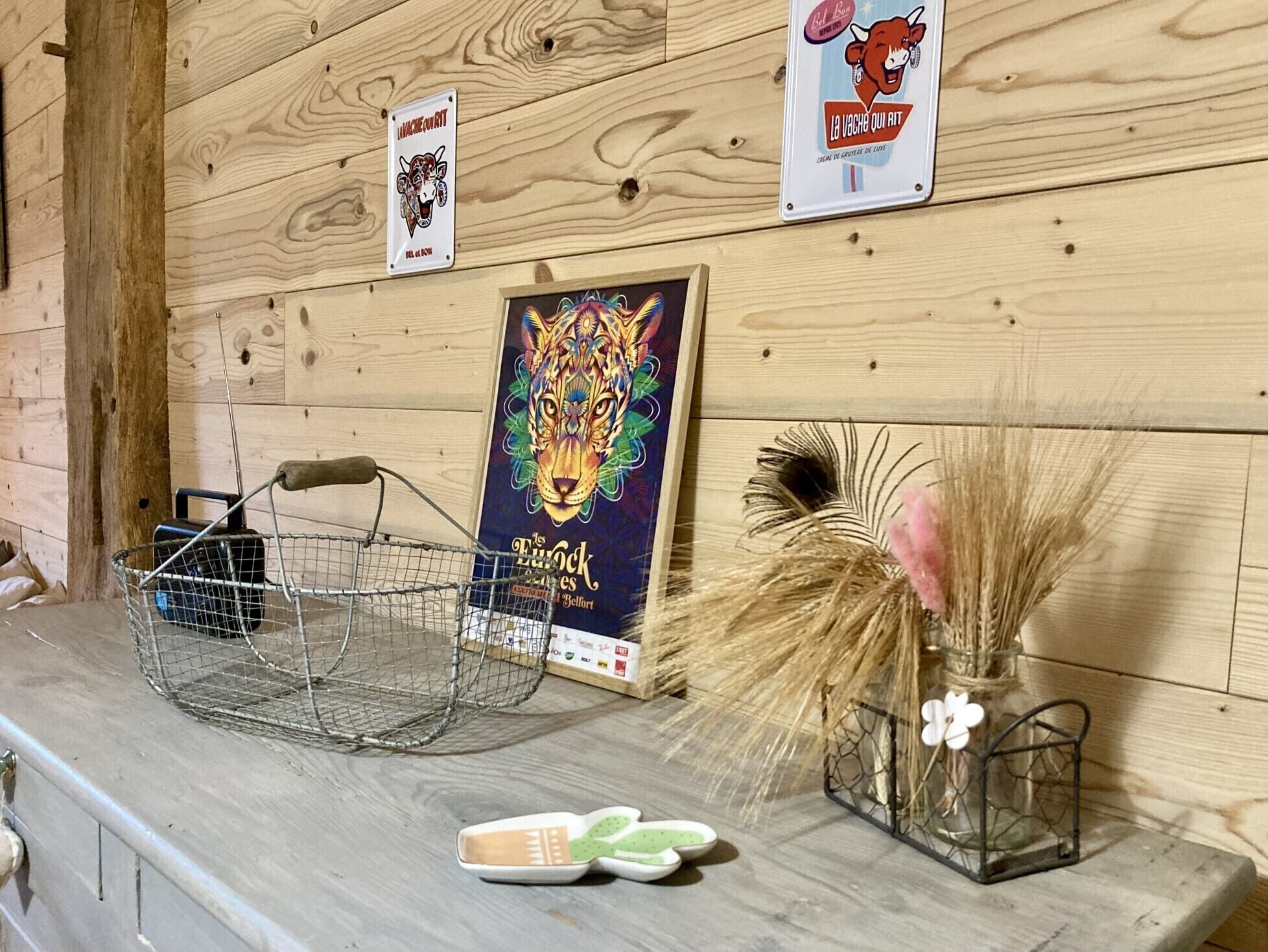 Un mur en bois orné d’une affiche encadrée, un panier en métal et un vase contenant des plantes séchées.