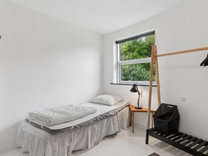 A minimalist bedroom with a single bed, a desk, a standing lamp, and a window with a view of greenery.
