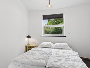 Een slaapkamer met een bed, nachtkastje, lamp en een raam met uitzicht op bomen.