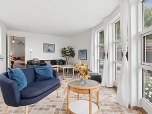 Een moderne woonkamer met een blauwe bank, een ronde houten tafel met een bloevas, en grote ramen met witte gordijnen.