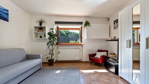 Un salon avec un canapé gris, un fauteuil rouge, une fenêtre donnant sur des arbres, un radiateur blanc et une étagère avec des plantes.
