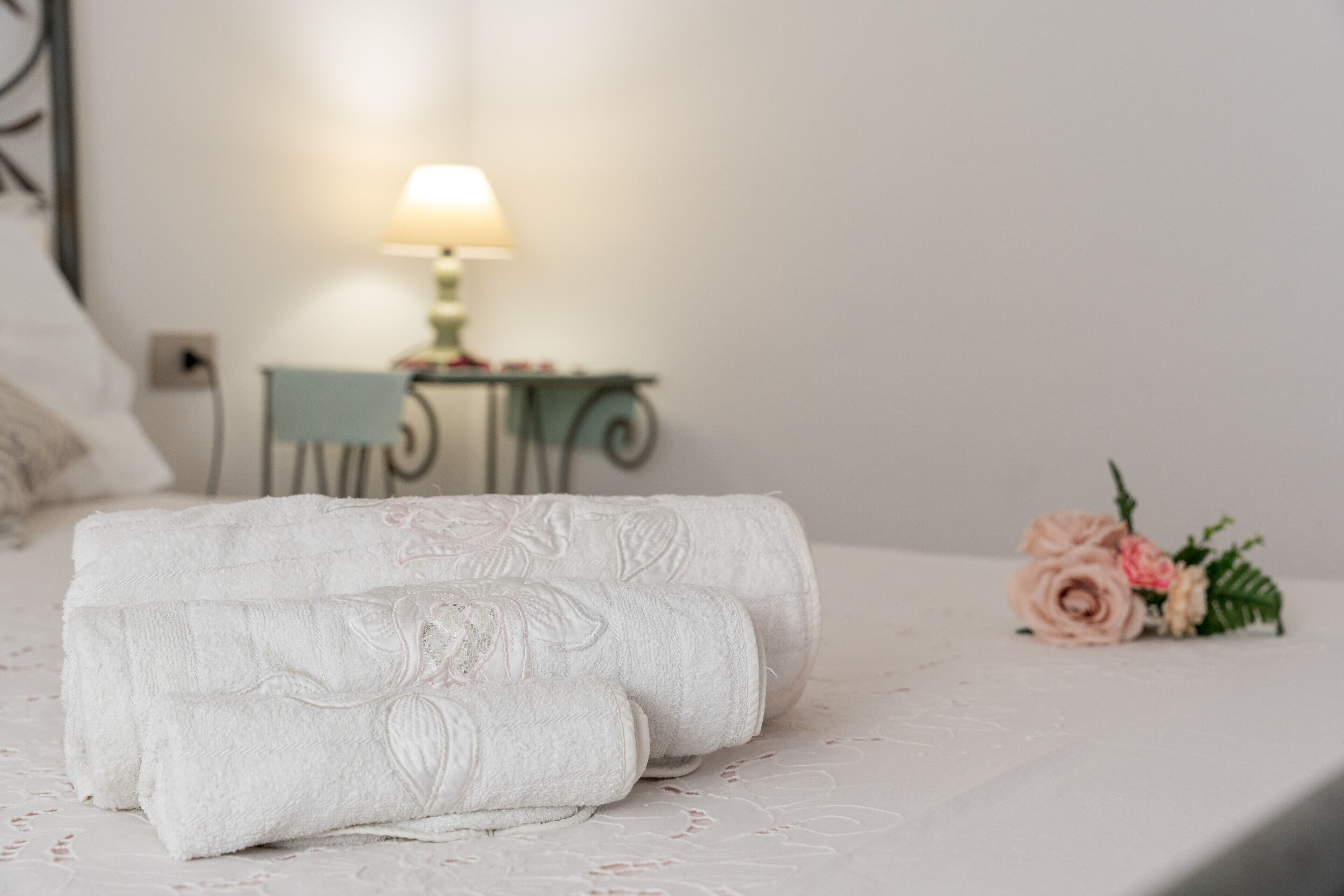 Un lit bien fait, avec des serviettes pliées en rouleaux et une petite table sur laquelle se trouvent une lampe et des fleurs.