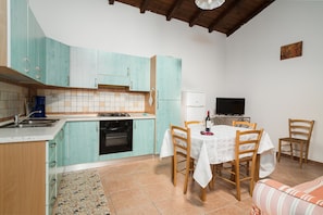Een keuken met blauwgroene kastjes, een eettafel met stoelen, een televisie en een koelkast.