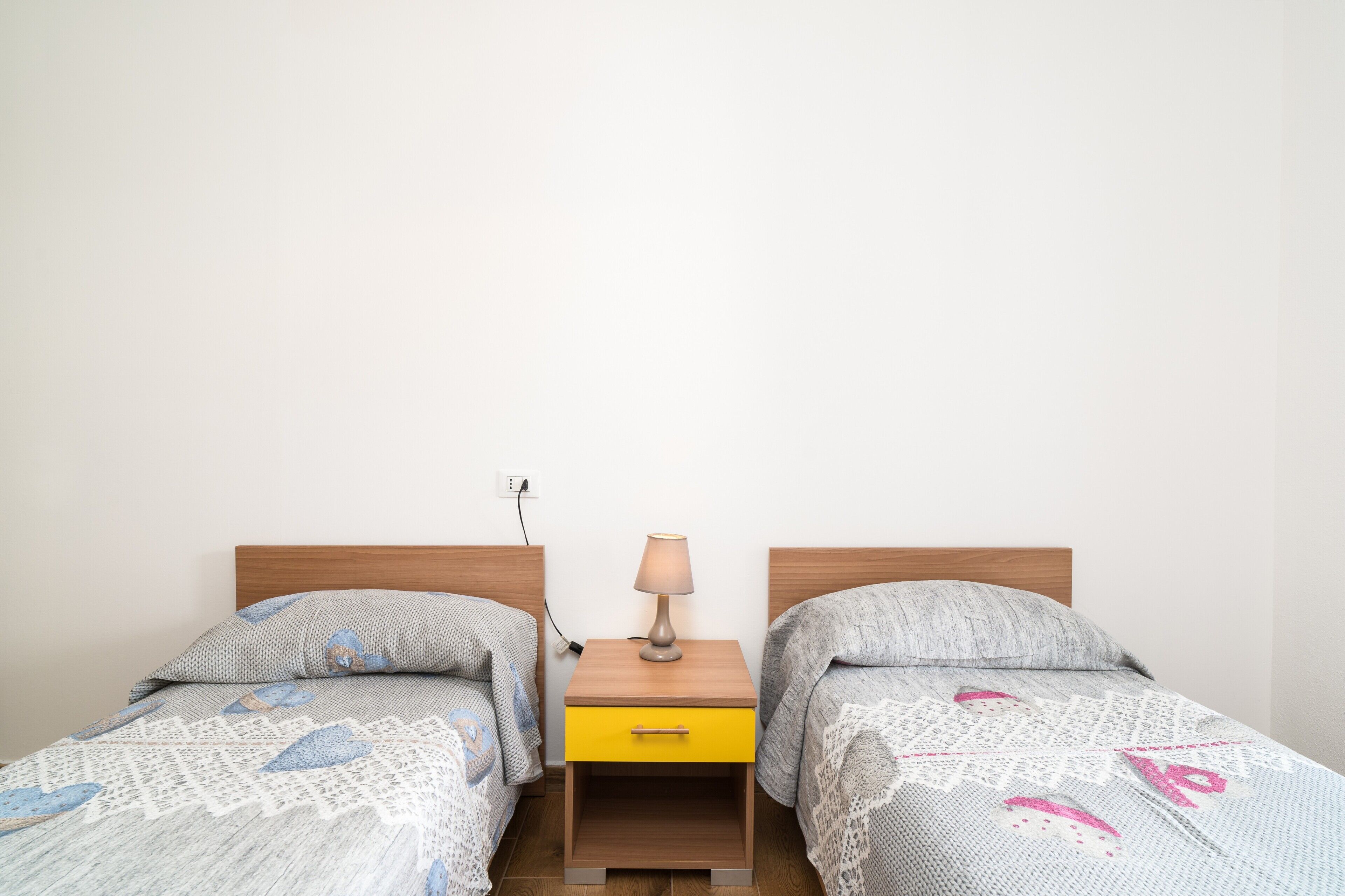 A room with two beds, a nightstand with a lamp, and a power outlet on the wall.