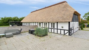 Een traditioneel gebouw met rieten dak, een gevel in het wit en zwart, een betegeld terras met tuinmeubilair en een tuin met hoge grassen.