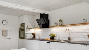 Een moderne keuken met witte kasten, een zwarte afzuigkap, een koffieapparaat en een spoelbak. Er hangt een klok aan de muur en staat een tafel met een witte mok en muffins.
