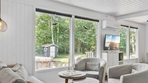 En moderne stue med sofa, lænestole og et lille bord. Store vinduer giver udsigt over en have og et skur.