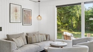 En moderne stue med en grå sofa, stribede puder, et rundt sofabord og et stort vindue med udsigt til en have.