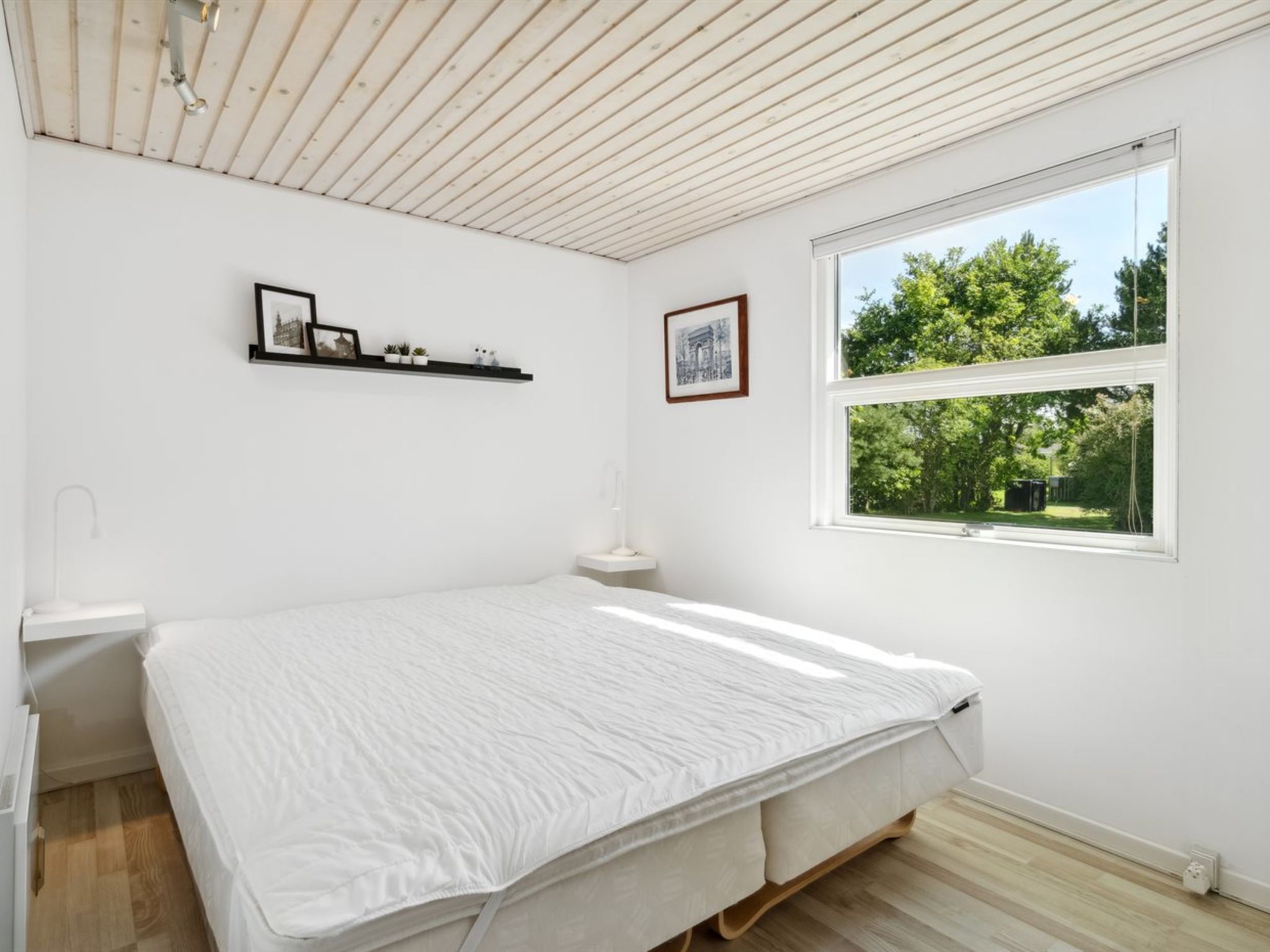 A bedroom with a large bed, a window with a view of greenery, a wall-mounted shelf with pictures, and a white ceiling.