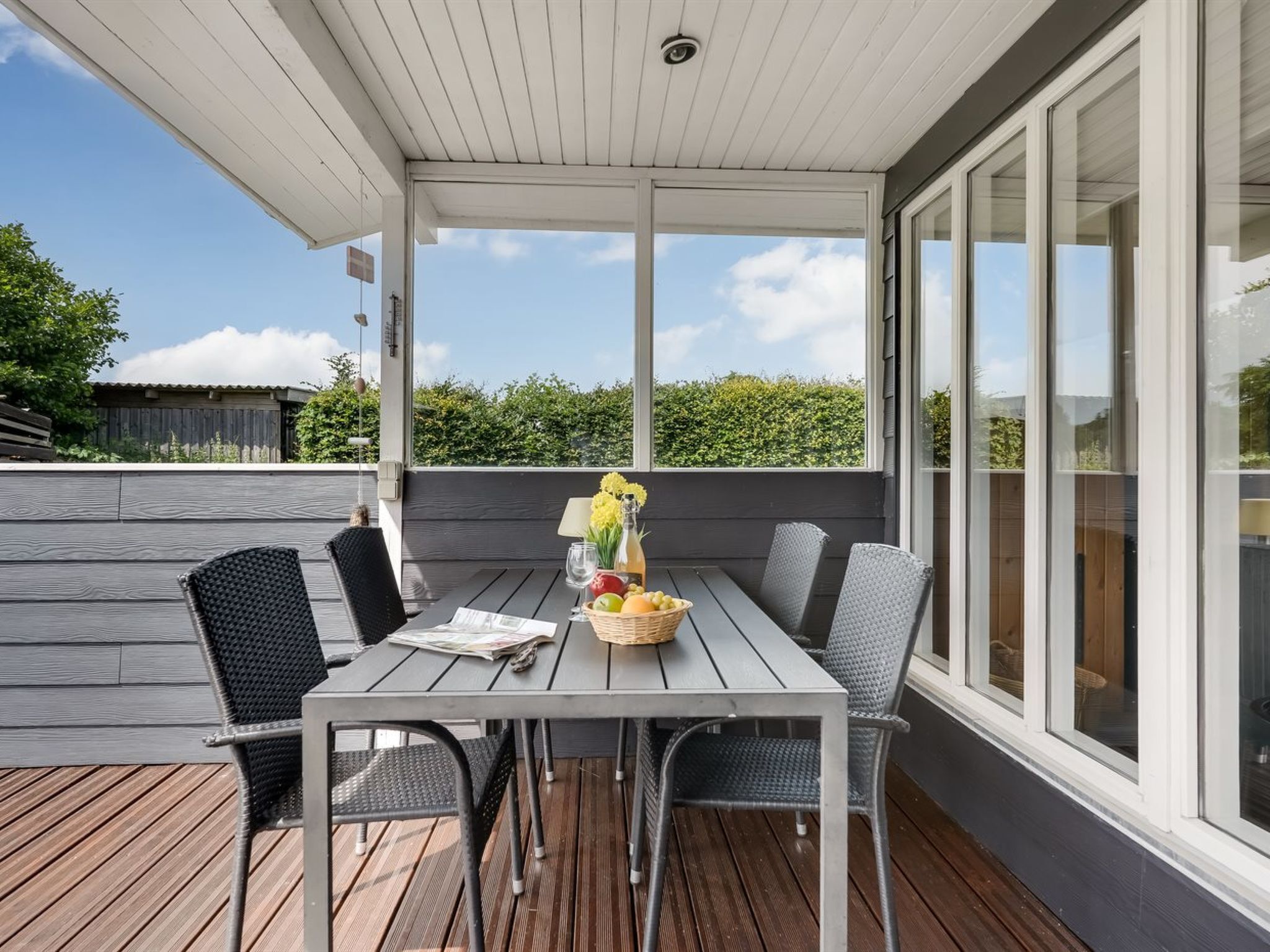 A covered outdoor seating area with a table set for two, surrounded by chairs, and a view of greenery.