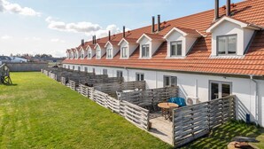 Eine Reihe weißer Gebäude mit roten Dächern und mehreren Schornsteinen, eine Holzterrasse mit Tisch und Stühlen sowie eine Rasenfläche mit einem Holzzaun.
