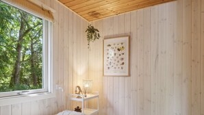 Un dormitorio con cama, ventana, planta colgante, mesita con lámpara y un cuadro en la pared.