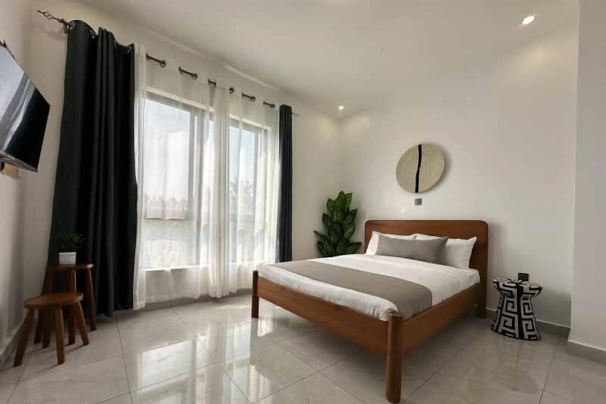 A modern bedroom with a wooden bed, a flat-screen TV mounted on the wall, a potted plant, and a round wall clock.