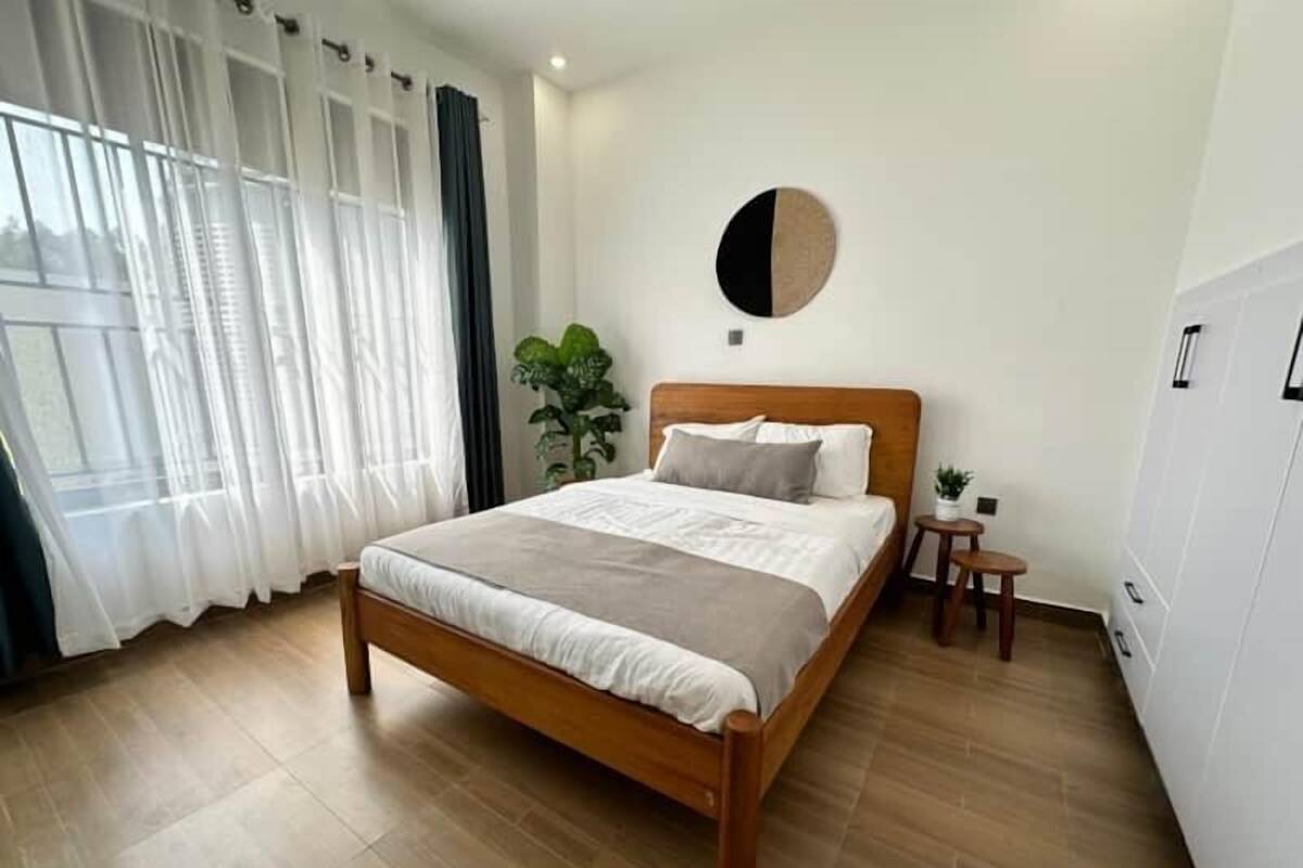 A bedroom with a wooden bed, white curtains, a round wall decoration, and a small plant.