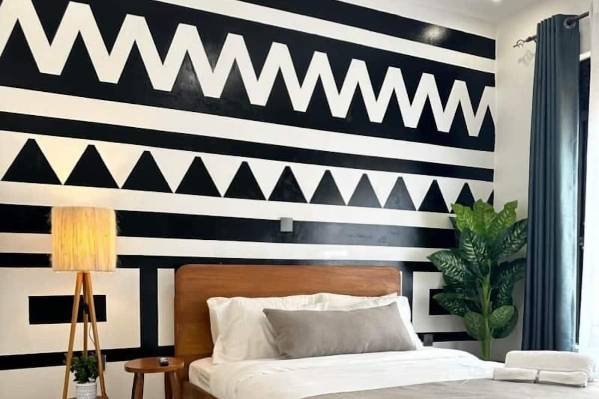 A modern bedroom with a wooden bed, a geometric patterned wall, a floor lamp, and a potted plant.